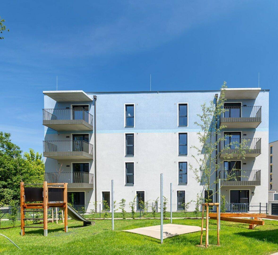 Modernes Wohngebäude mit blauer und weißer Fassade, Balkonen und einem Spielplatz im Vordergrund.