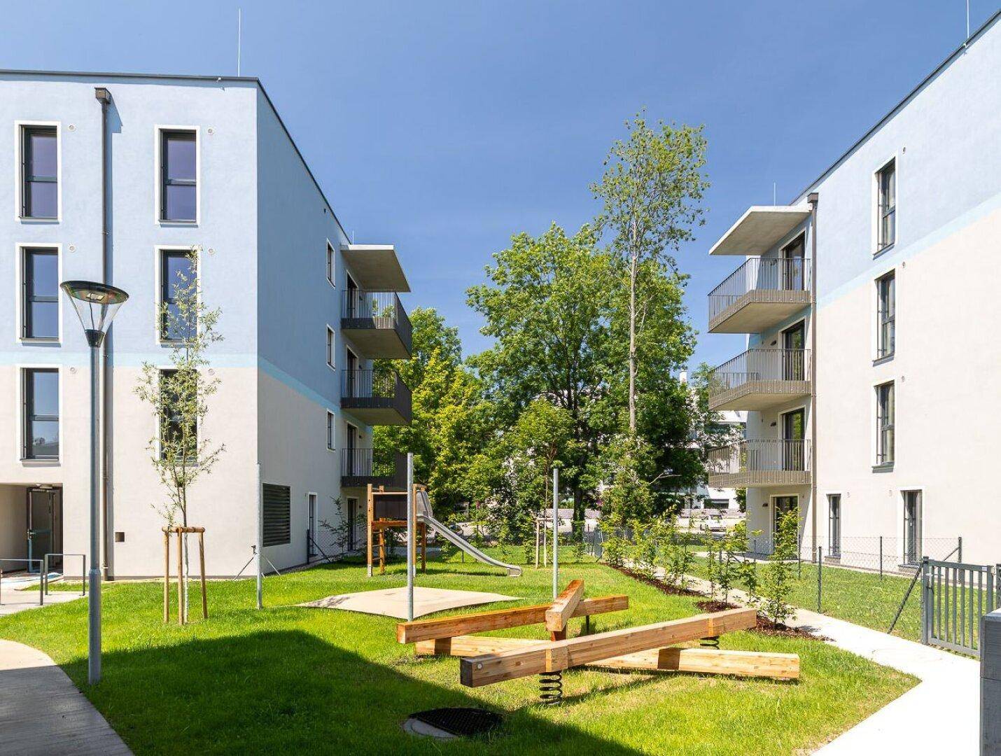Kinderspielplatz mit Schaukel und Rutsche zwischen modernen Wohngebäuden mit blauer Fassade.