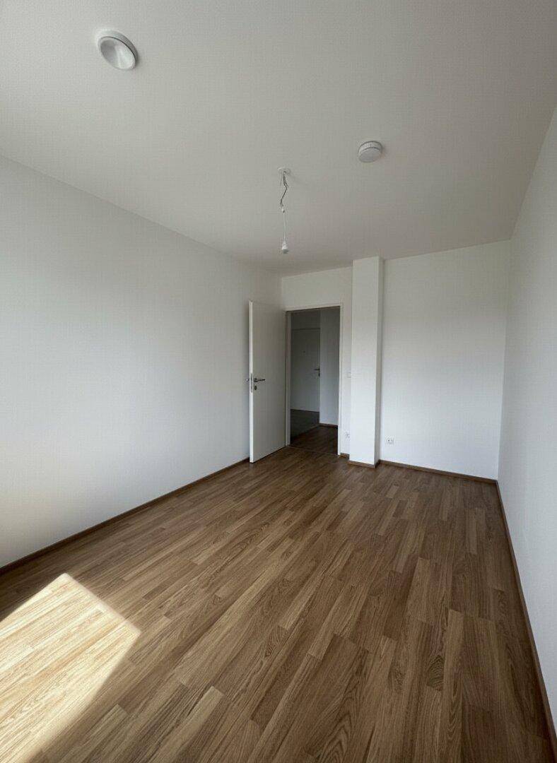 Helles, leeres Zimmer mit Laminatboden und integrierten Deckenleuchten.