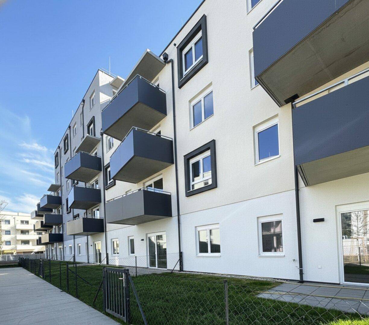 Moderne Wohnanlage mit weißer Fassade, dunklen Balkonen und kleinen Grünflächen im Erdgeschoss.