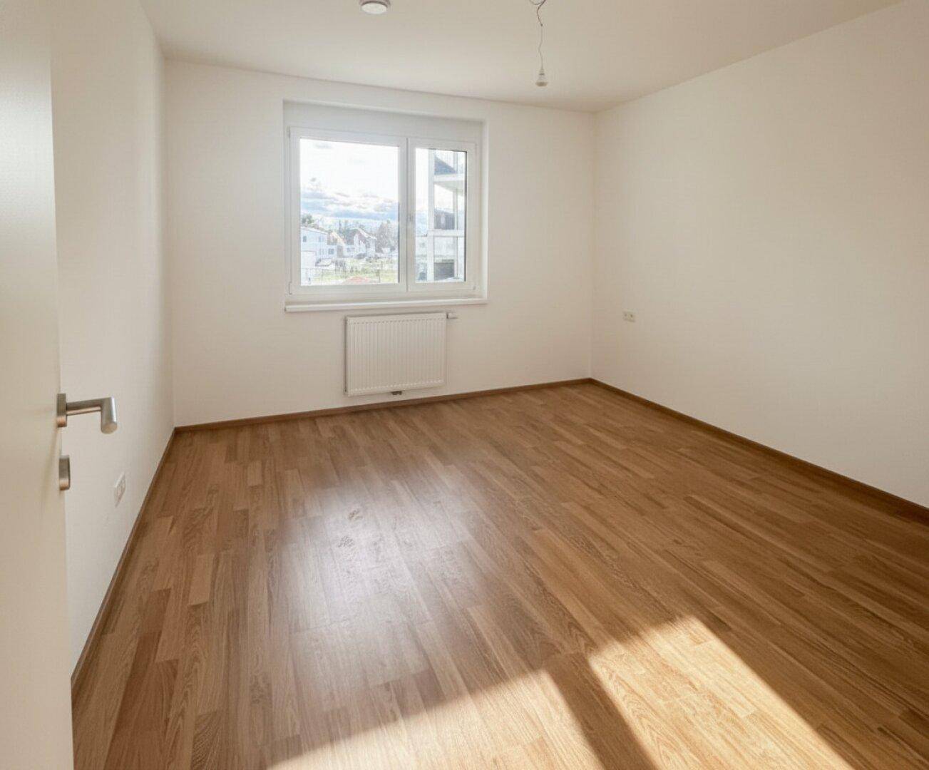 Helles, leeres Zimmer mit Holzboden und großem Fenster, das einen weiten Blick bietet.
