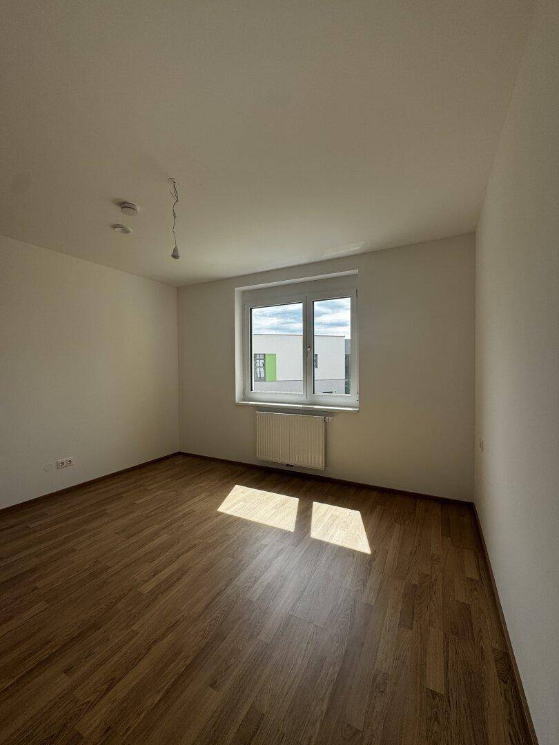 Leeres Zimmer mit Holzlaminatboden und einem Fenster, das viel natürliches Licht hereinlässt.