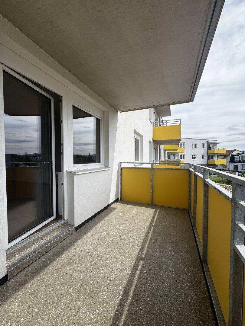Heller Balkon mit Betonboden und gelben Akzenten, bietet Zugang zu einem modernen Wohngebäude.