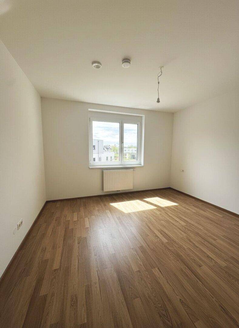 Helles Schlafzimmer mit Laminatboden und großem Fenster, das viel Tageslicht hereinlässt.