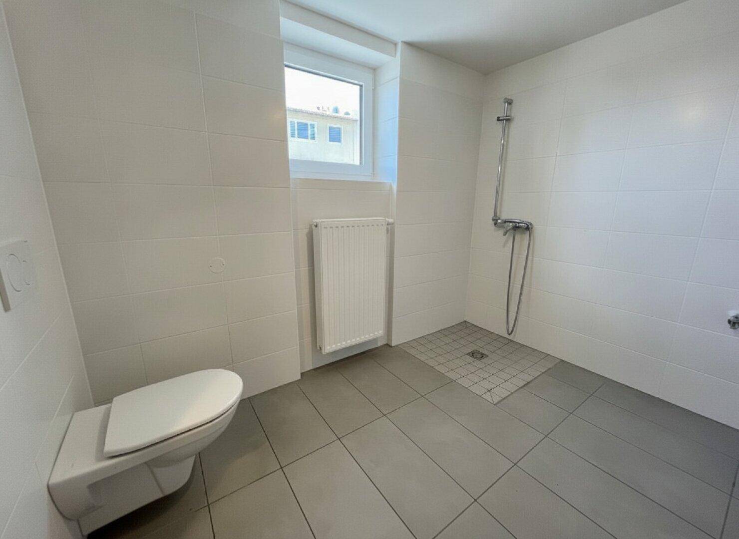 Modernes Badezimmer mit weißen Wandfliesen, grauen Bodenfliesen, bodengleicher Dusche und Fenster.
