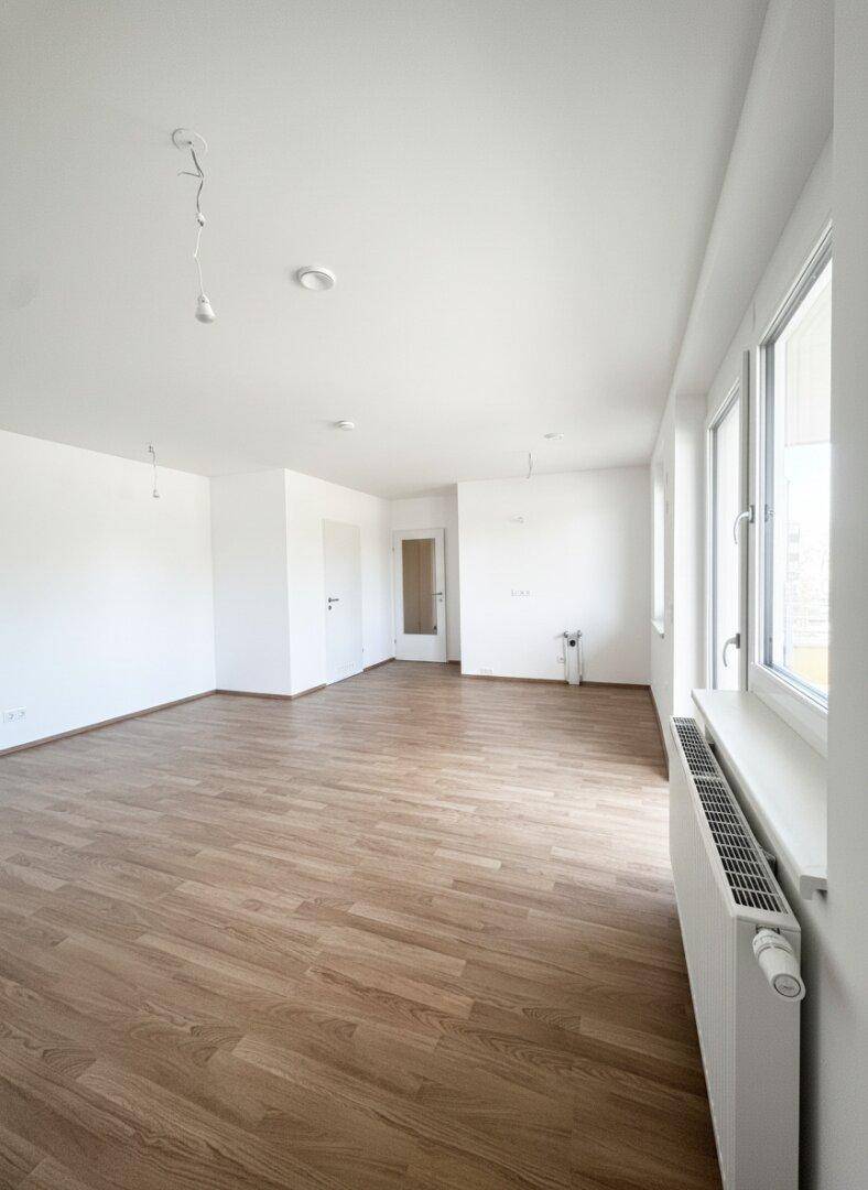 Unmöblierter Wohnbereich mit Laminatboden, weißen Wänden und Heizkörper unter dem Fenster.