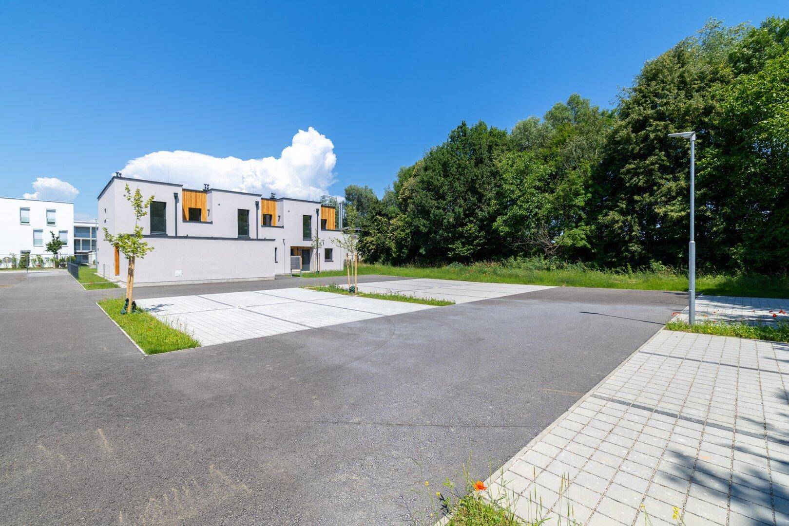Weitläufiger Parkplatz mit Bäumen und modernen Reihenhäusern im Hintergrund unter blauem Himmel.
