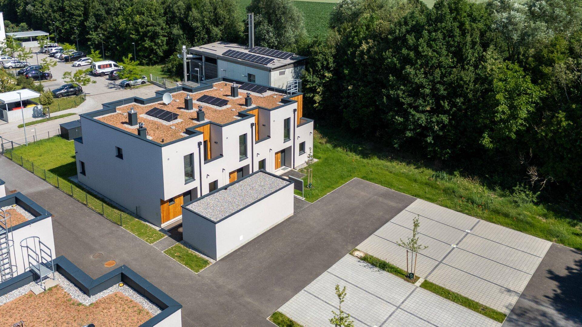 Luftaufnahme einer Gruppe moderner Reihenhäuser mit Flachdächern und Photovoltaikanlagen, umgeben von Natur.