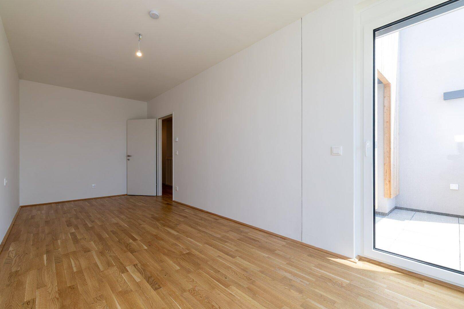 Helles, leeres Zimmer mit Eichenparkettboden und Zugang zu einem kleinen Balkon.