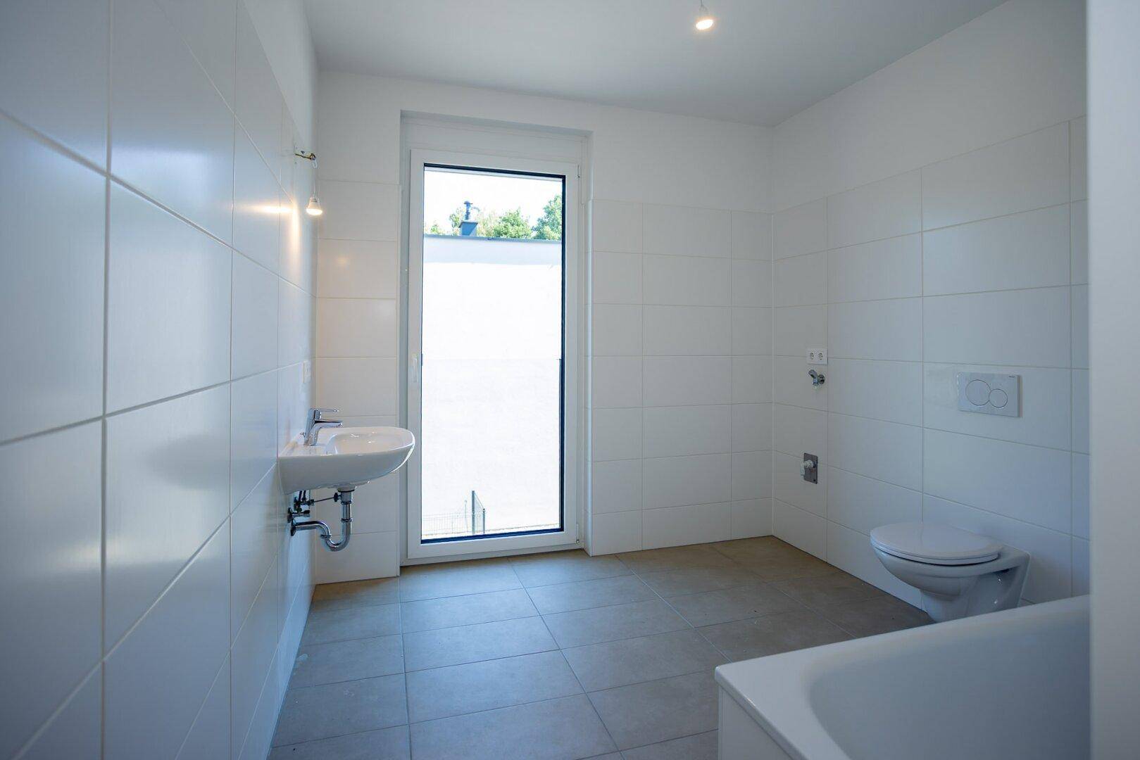 Helles Badezimmer mit Fenster, Waschbecken, Badewanne und weißen Wandfliesen.
