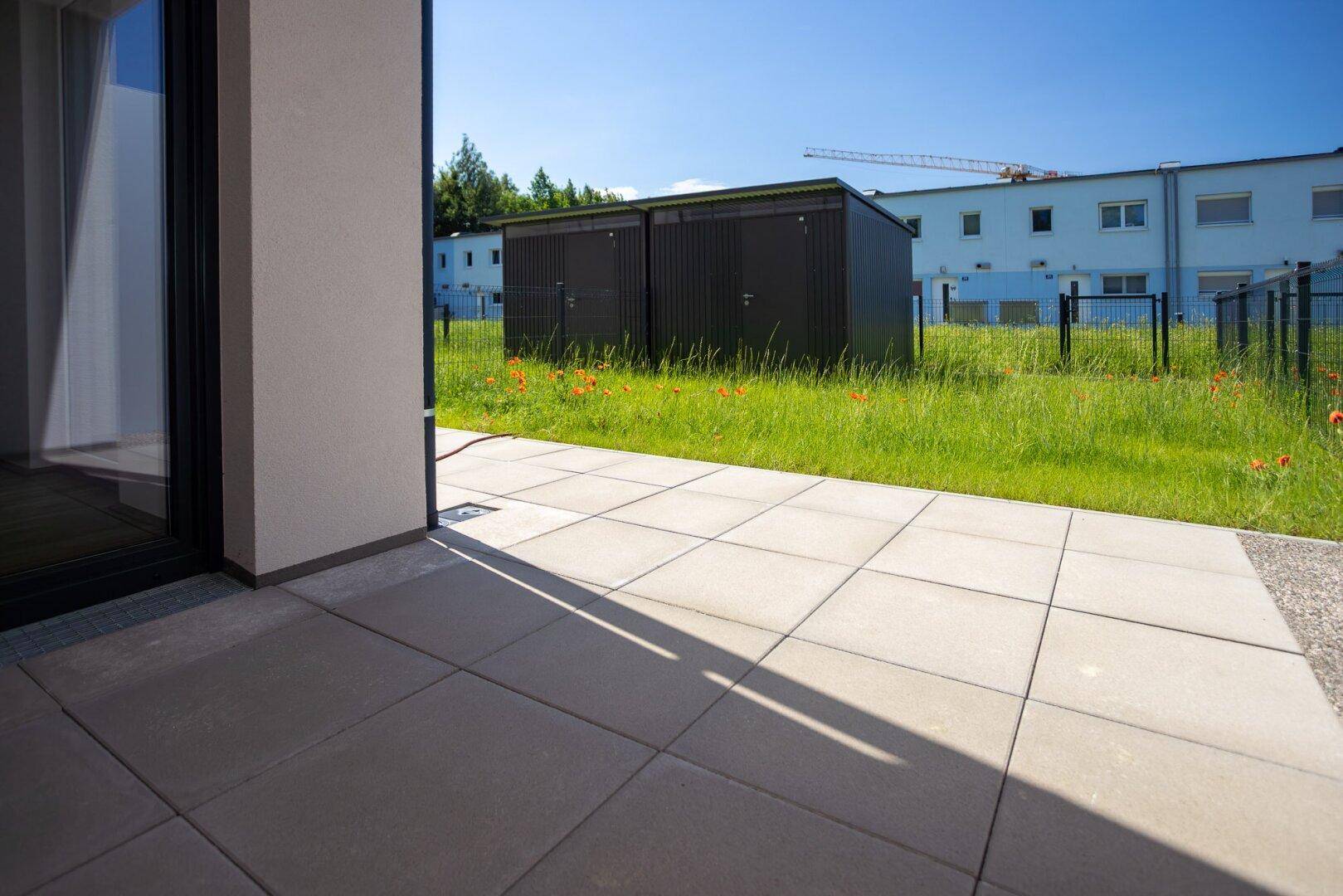 Gepflasterte Terrasse mit Blick auf den grünen Garten und zwei Gartenhäuser unter blauem Himmel.