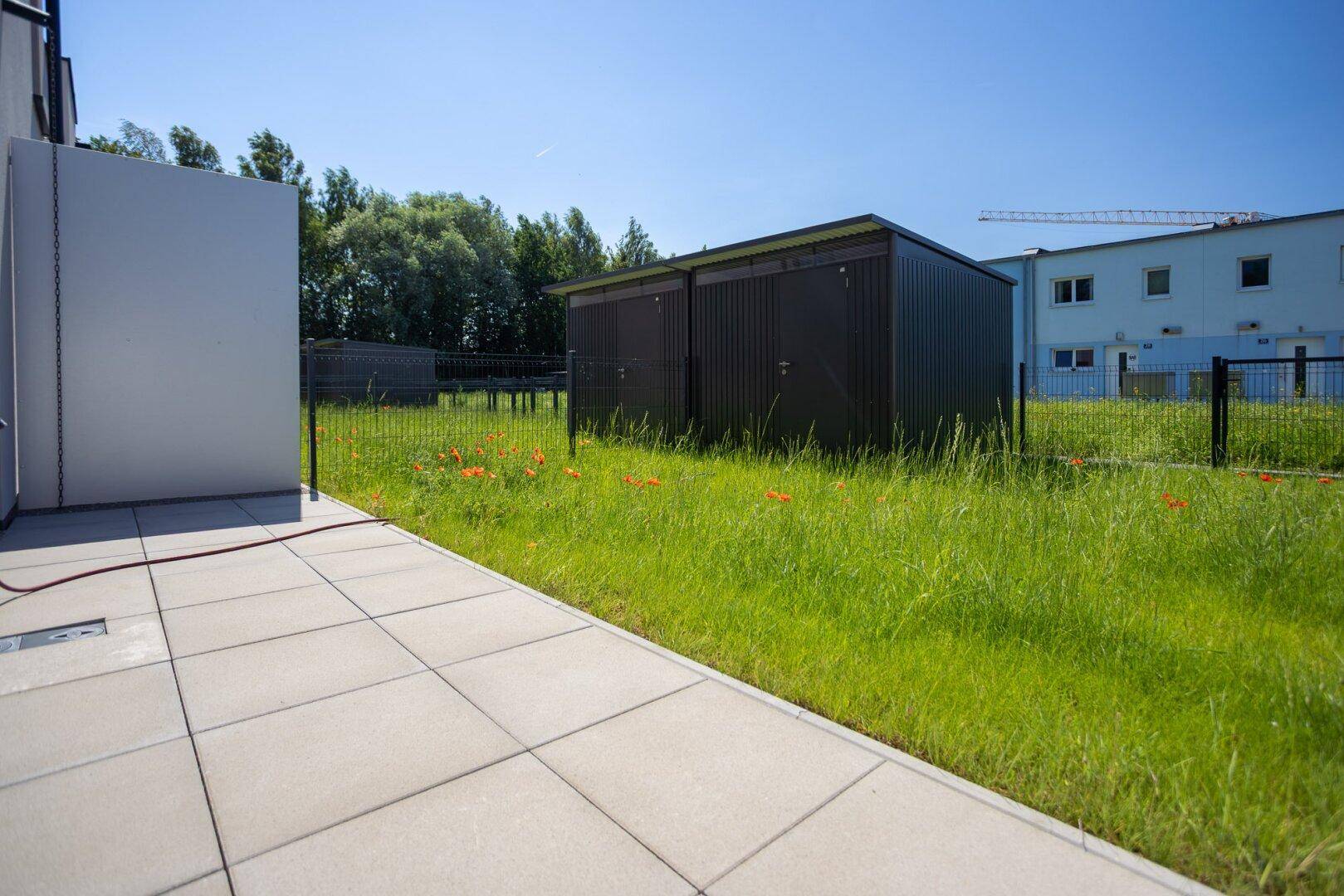 Weitläufige Terrasse mit angrenzendem Rasenbereich und praktischen Gartenhäusern im Hintergrund.