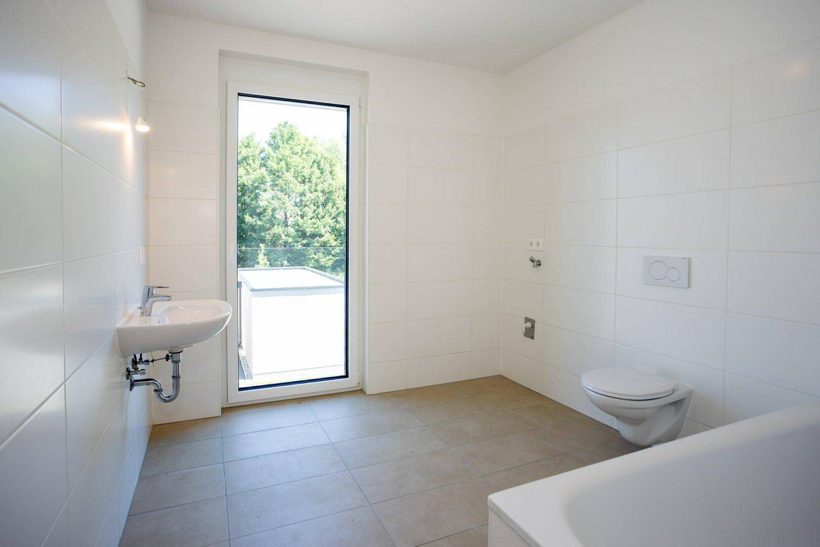 Helles Badezimmer mit weißen Fliesen, Waschbecken, WC und großem Fenster für viel Tageslicht.