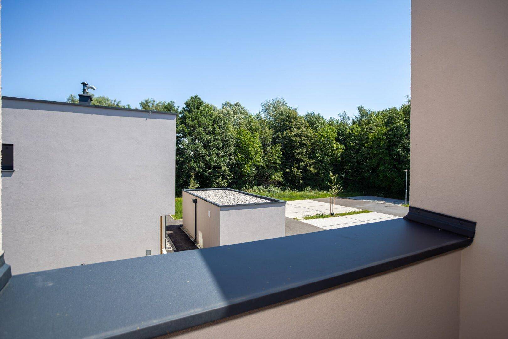 Blick vom Balkon auf einen gepflasterten Bereich und eine Reihe von Bäumen unter klarem Himmel.