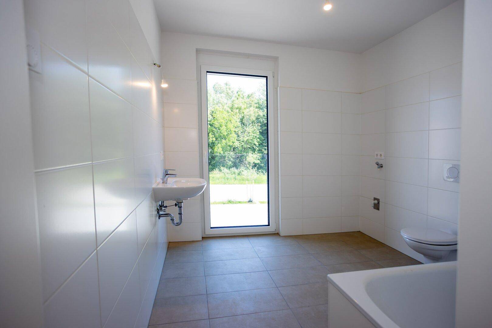 Helles Badezimmer mit Badewanne, wandhängendem WC, Waschbecken und großem Fenster für Tageslicht.