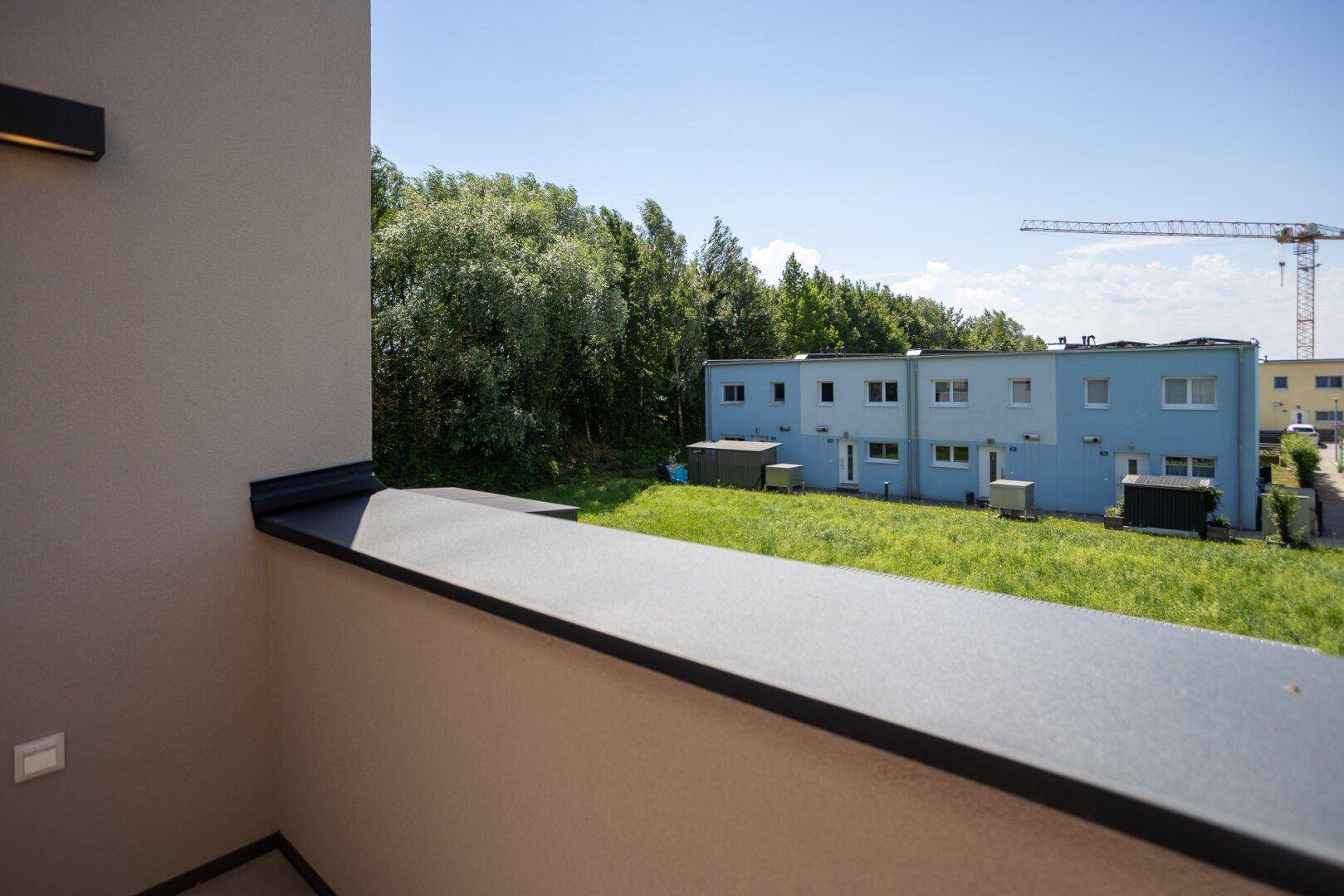 Seitlicher Blick vom Balkon auf die grüne Umgebung und benachbarte Häuser mit einem Kran im Hintergrund.