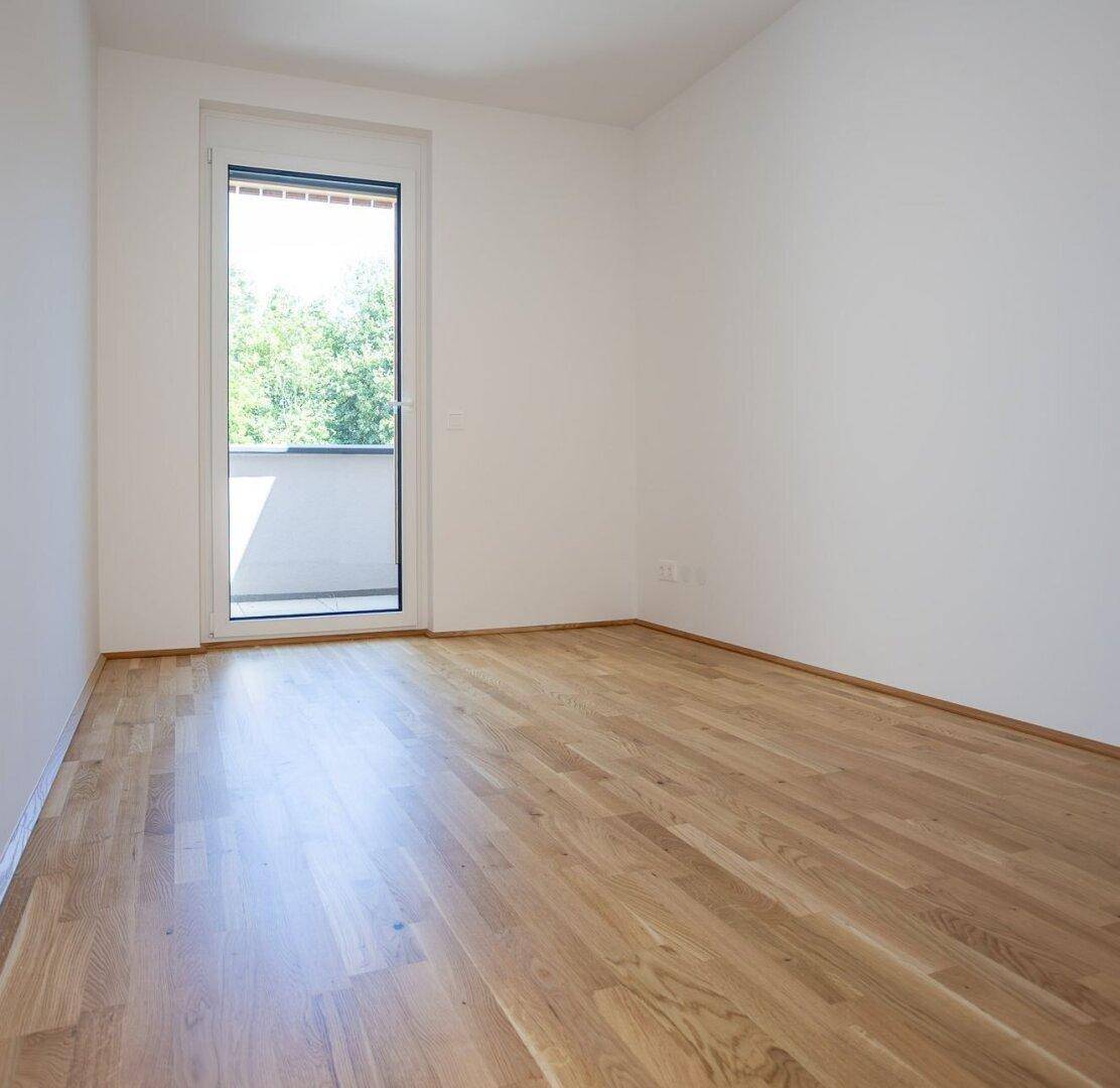 Helles Schlafzimmer mit Parkettboden und Zugang zu einem französischen Balkon mit Grünblick.