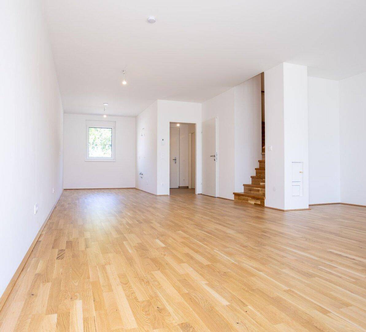 Geräumiger Wohnbereich mit Parkettboden, einem Fenster und Zugang zu einer Holztreppe.