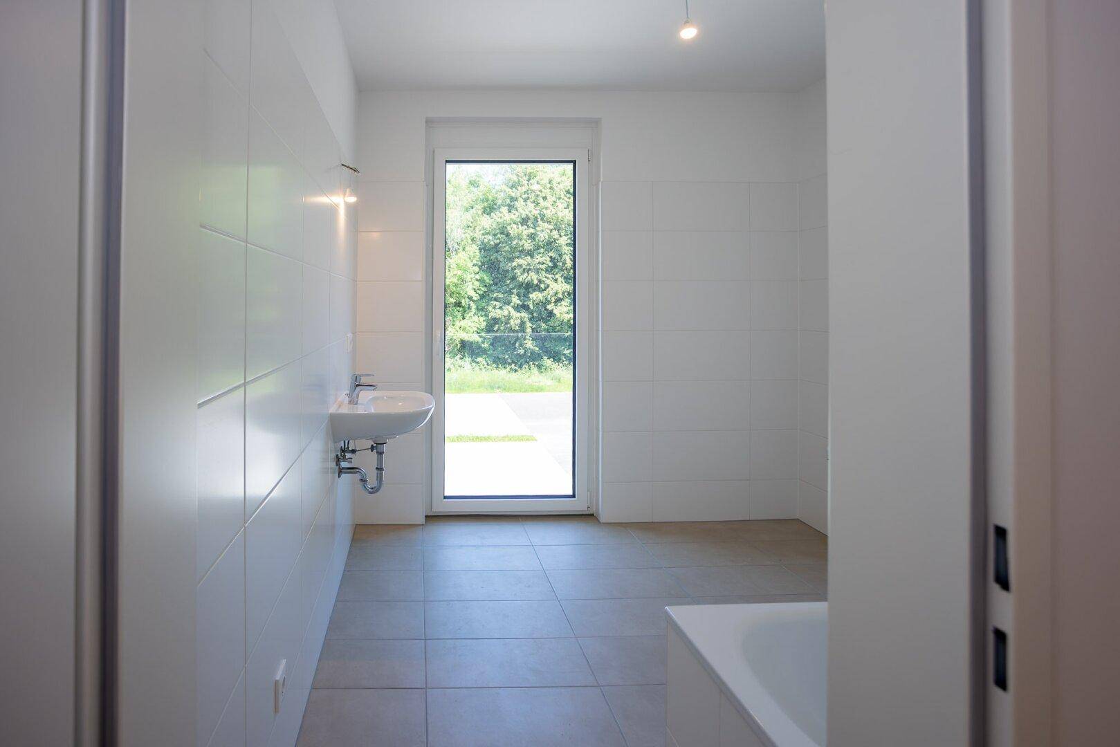 Modernes Badezimmer mit weißen Fliesen, einem Waschbecken und einem großen Fenster mit Grünblick.