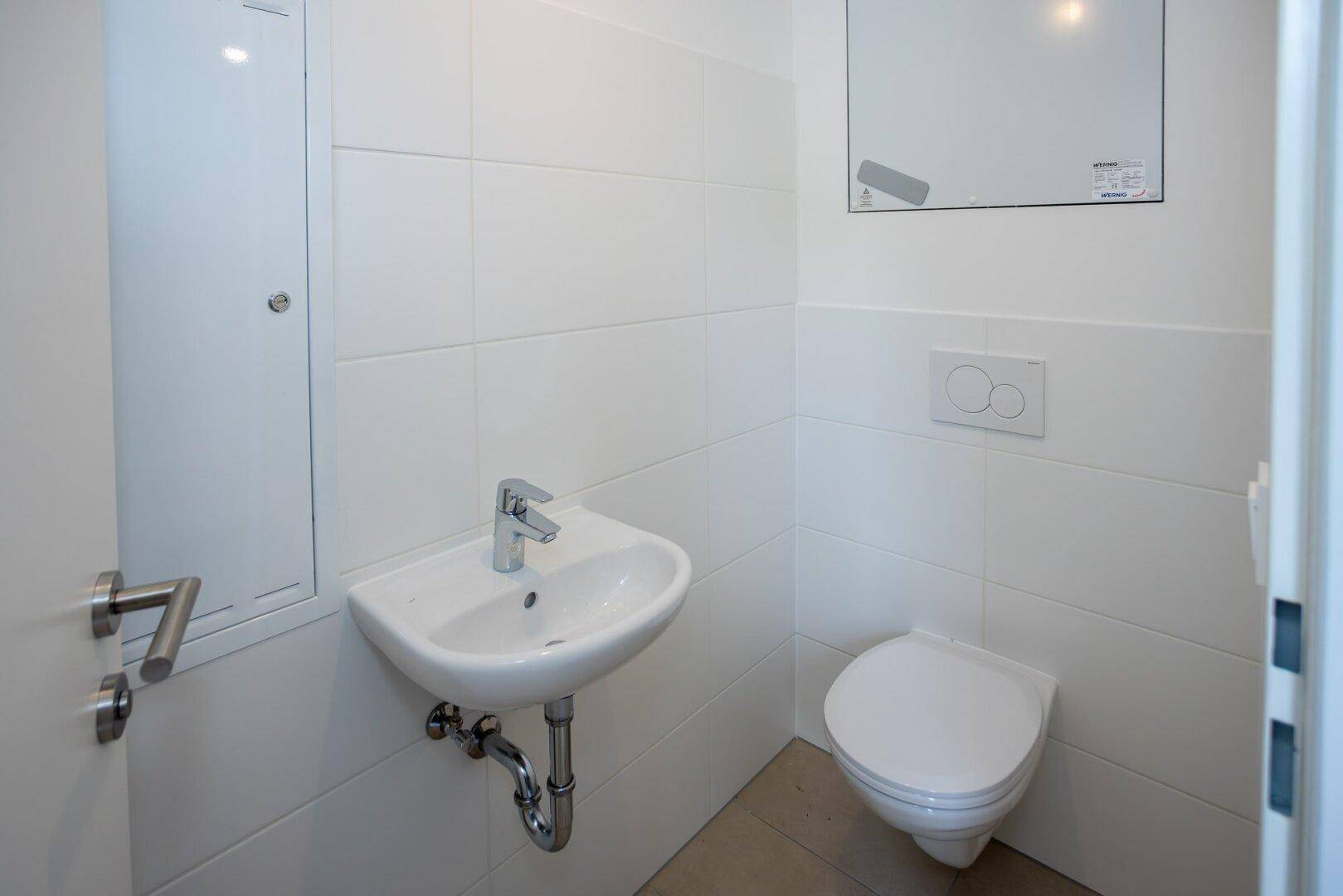 Modernes Gäste-WC mit weißen Fliesen, wandhängendem WC und kleinem Handwaschbecken.