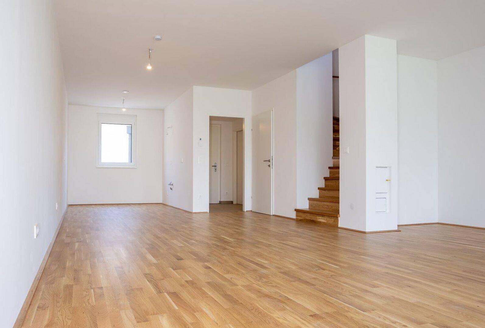 Heller Wohnbereich mit Eichenparkett, weißen Wänden und einer Holztreppe ins Obergeschoss.