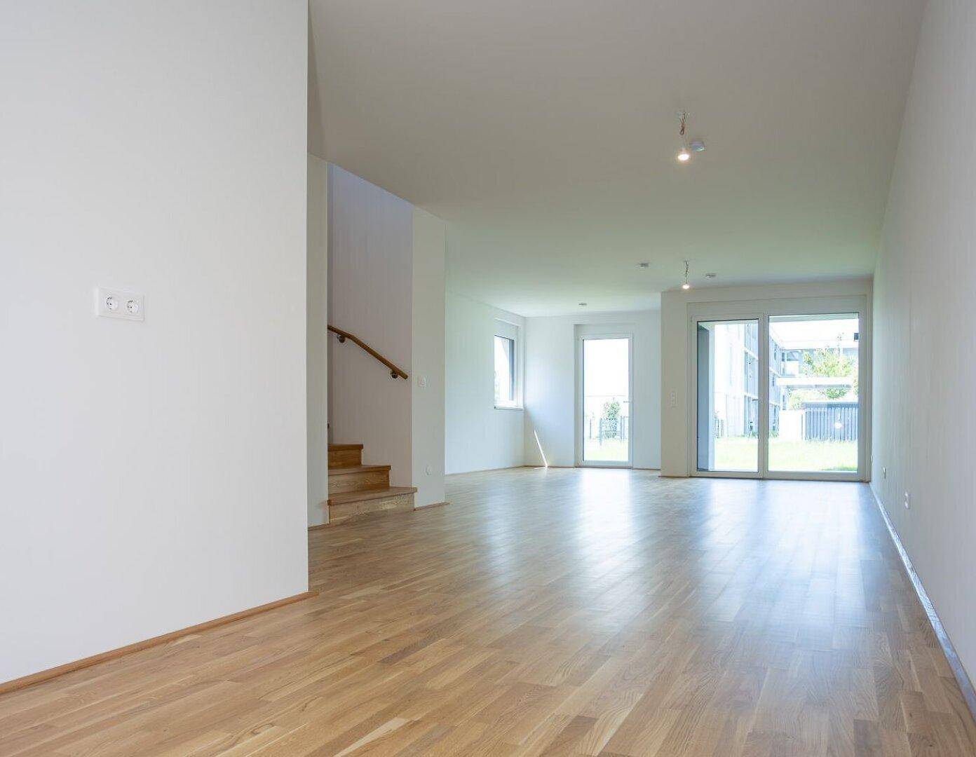 Heller, leerer Wohnbereich mit Eichenparkettboden, weißen Wänden und Zugang zum Garten.