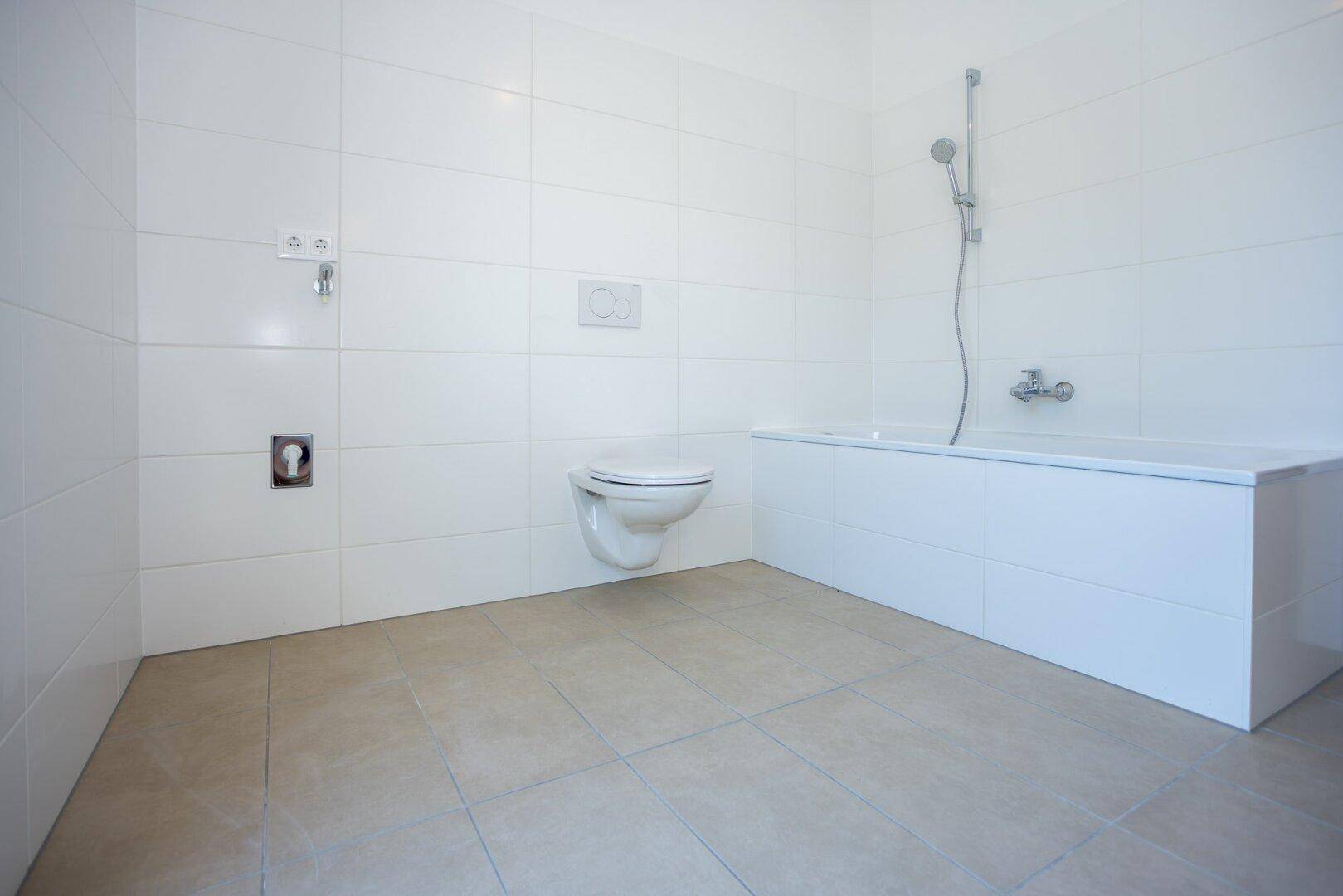 Helles Badezimmer mit weißen Wandfliesen, Badewanne mit Duschfunktion und wandhängendem WC.
