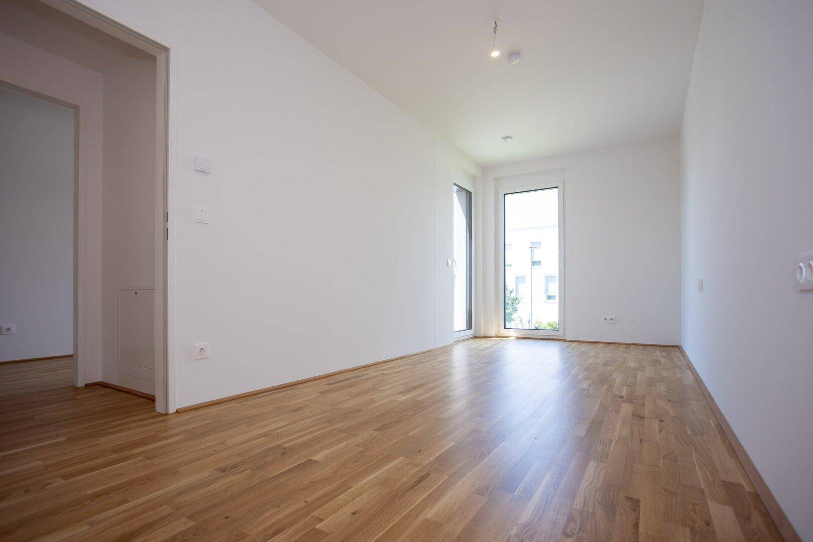 Leeres Schlafzimmer mit hellem Parkettboden, weißen Wänden und Zugang zu einem Balkon.