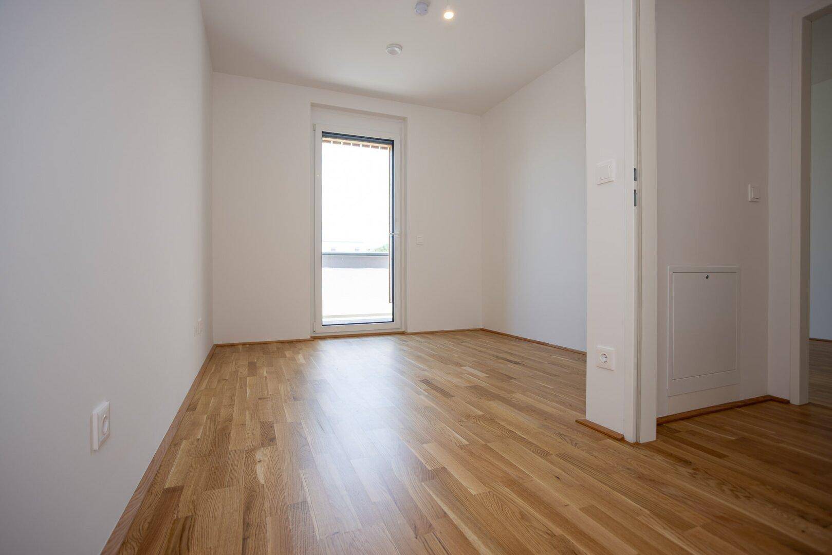 Helles, leeres Zimmer mit Eichenparkettboden, weißen Wänden und einem großen Fenster.