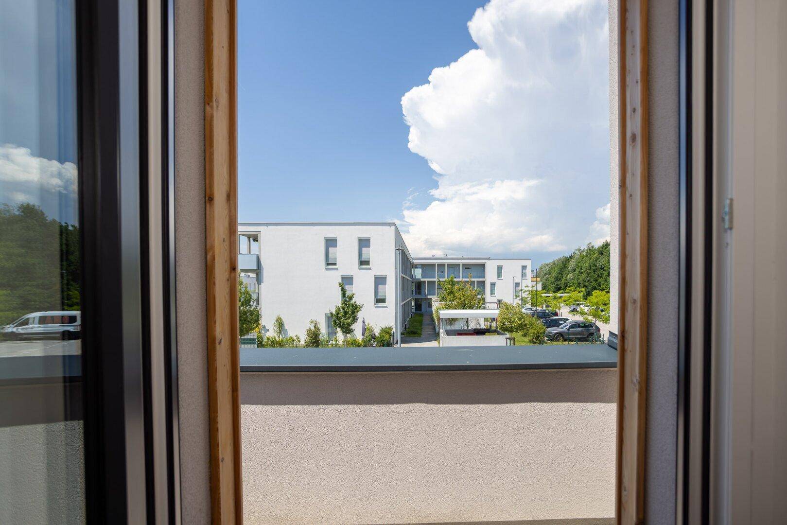 Blick aus dem Zimmer durch die offene Balkontür auf die umliegenden Gebäude und Bäume.