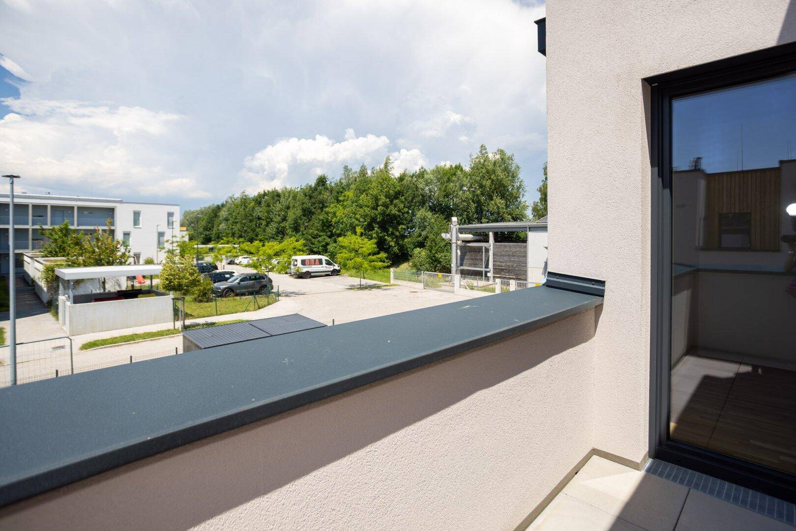Blick vom Balkon auf den Parkplatz und die umliegende grüne Landschaft mit Bäumen.