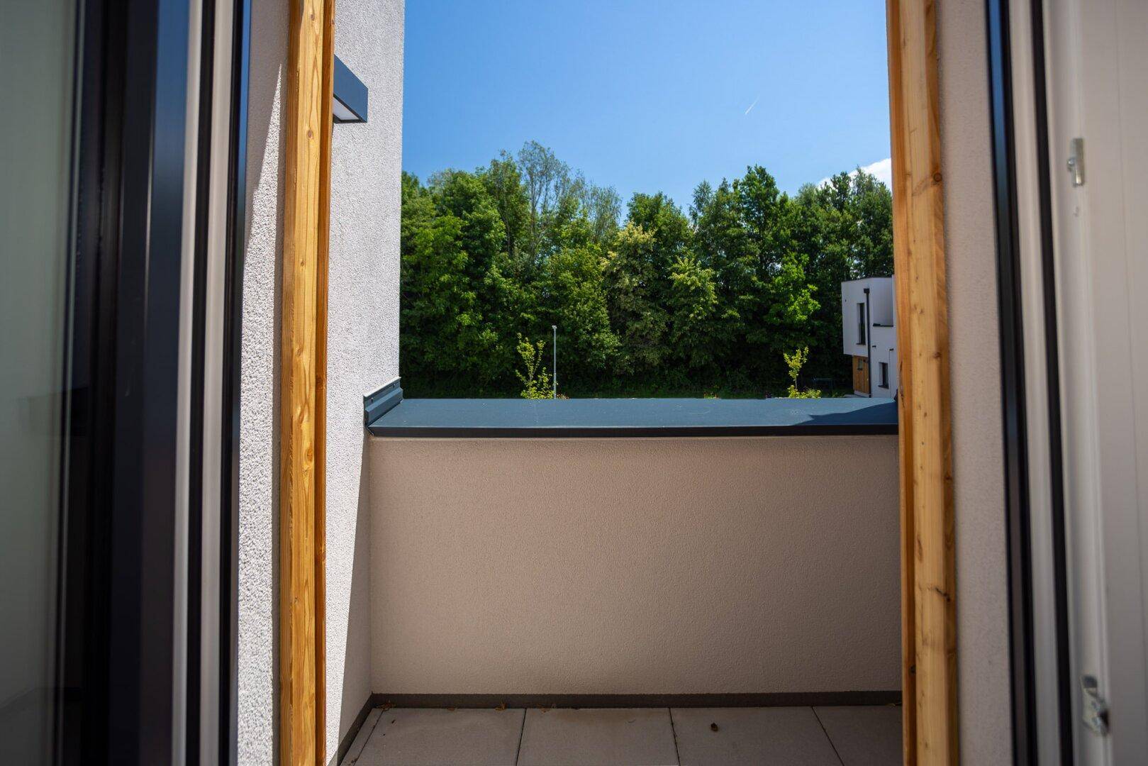 Blick aus dem Zimmer auf den Balkon und die grüne Umgebung mit vielen Bäumen.