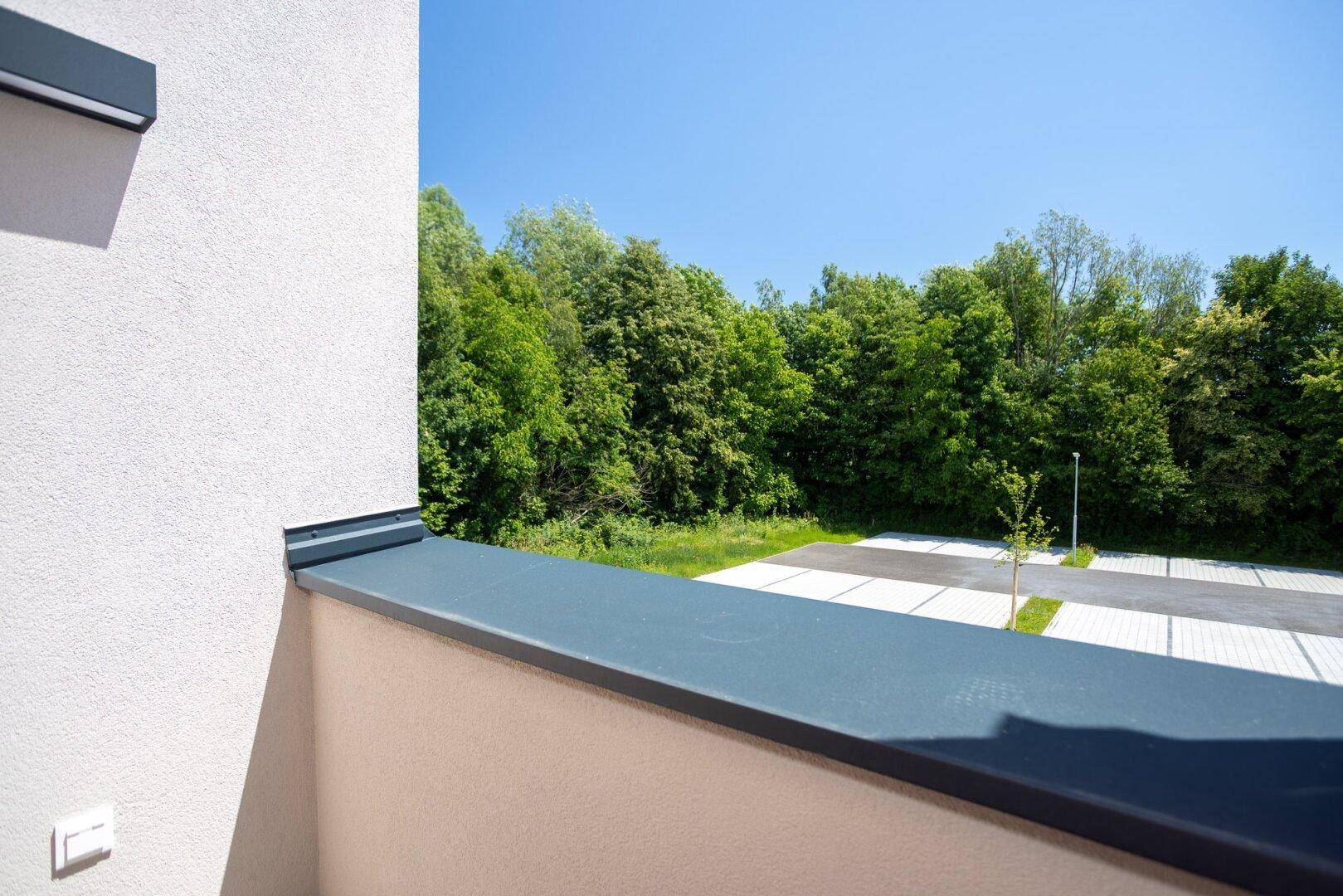Aussicht vom Balkon auf einen Parkplatz und eine dichte Baumreihe im Hintergrund.
