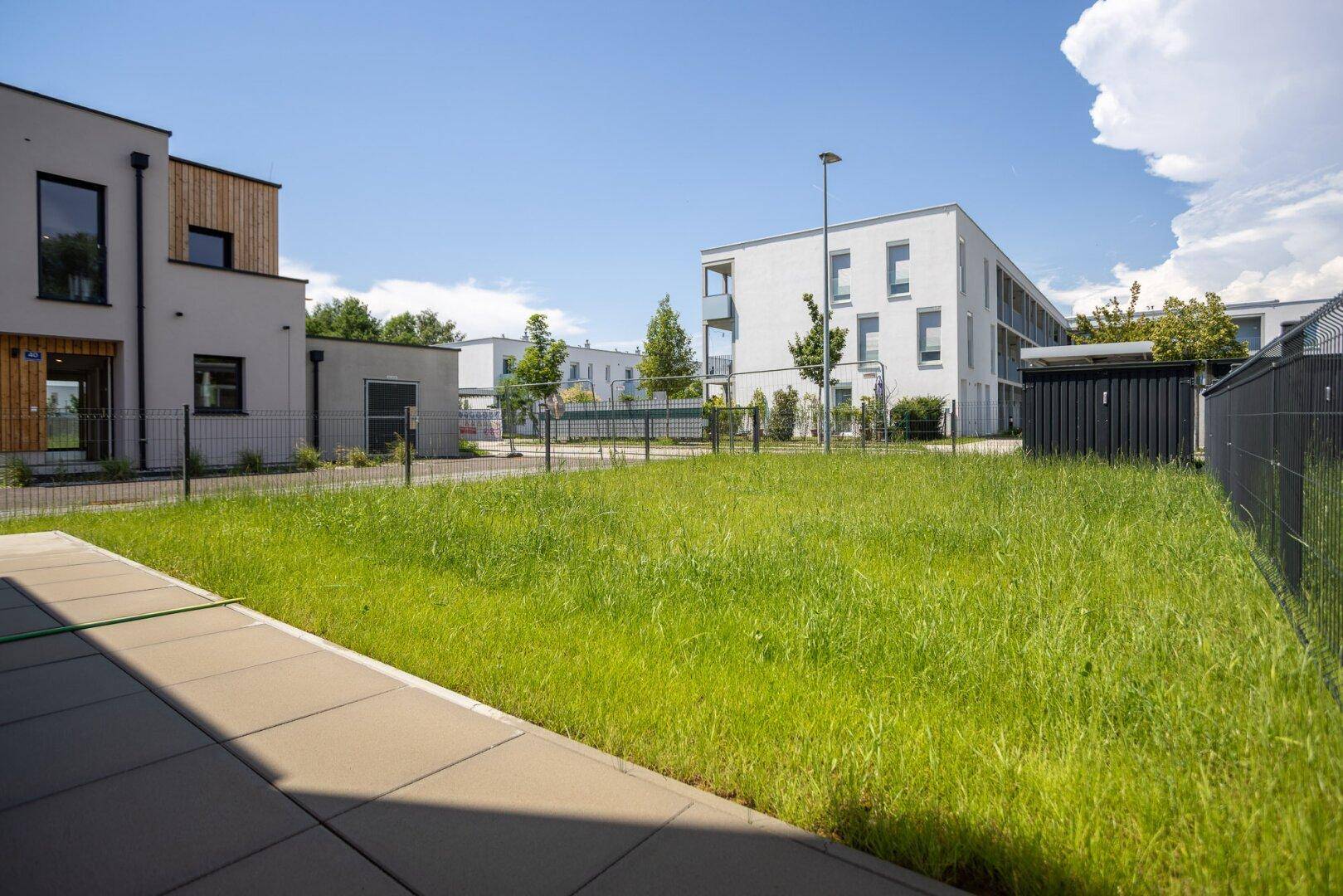 Grüne Rasenfläche mit gepflastertem Weg und modernen Wohngebäuden im Hintergrund.