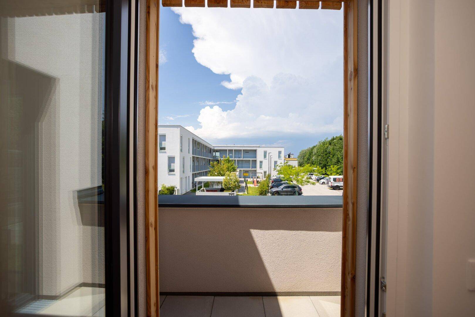 Blick durch die Balkontür auf die Wohnanlage und den blauen Himmel mit weißen Wolken.