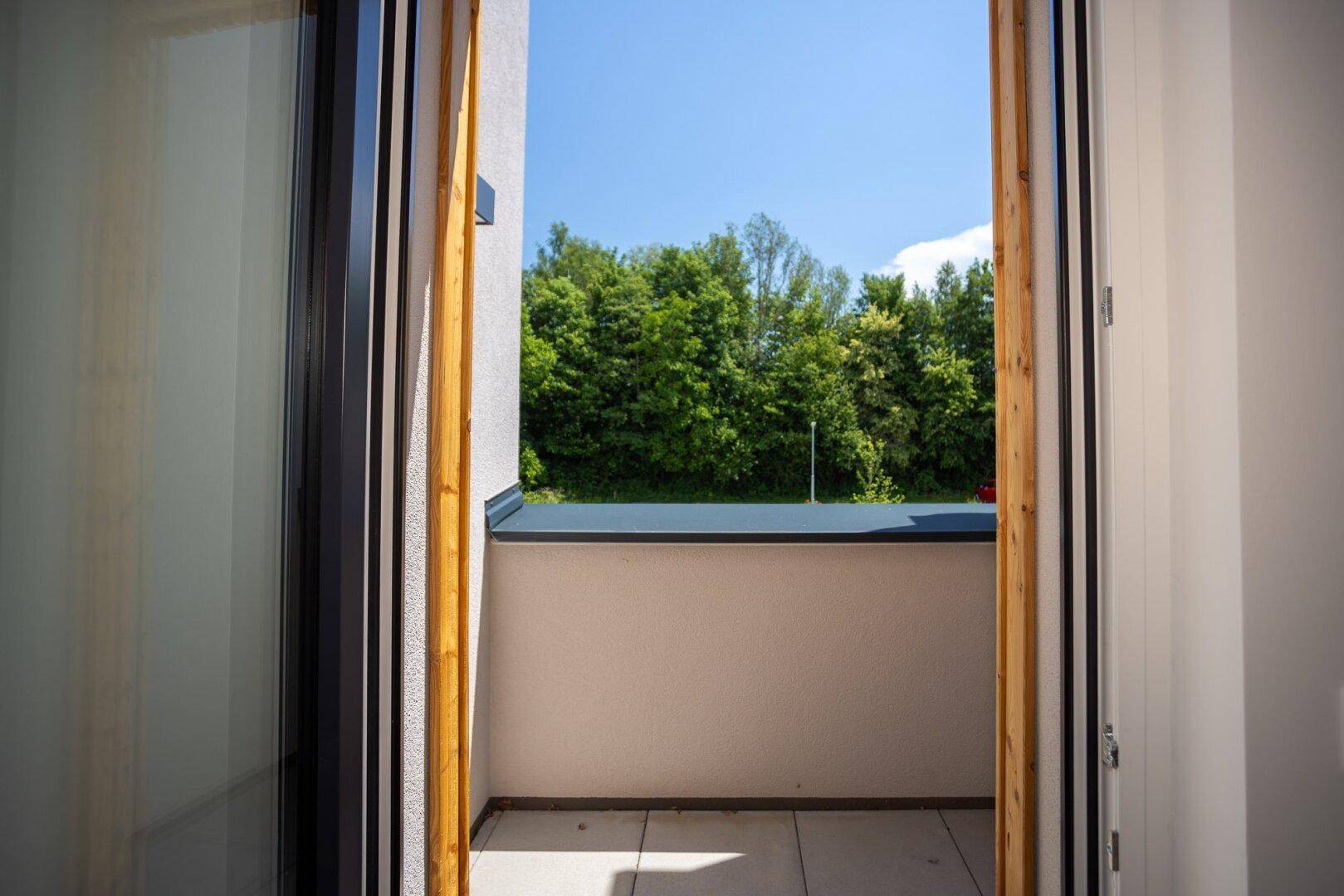 Aussicht durch die Balkontür auf eine grüne Baumreihe und den klaren blauen Himmel.