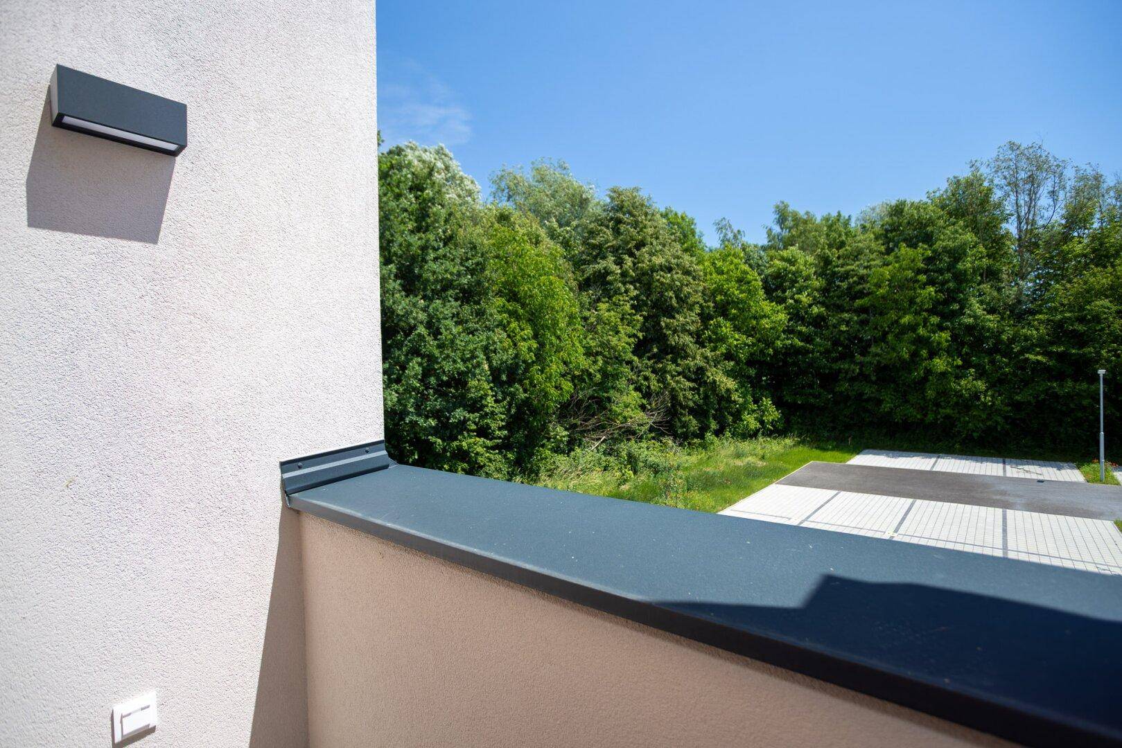 Blick vom Balkon auf eine grüne Baumreihe und einen Parkplatz unter strahlend blauem Himmel.