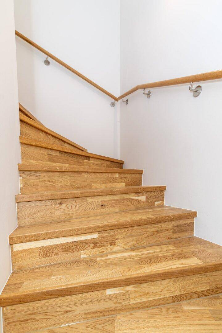 Holztreppe mit hellem Geländer und weiß gestrichenen Wänden, die in die obere Etage führt.