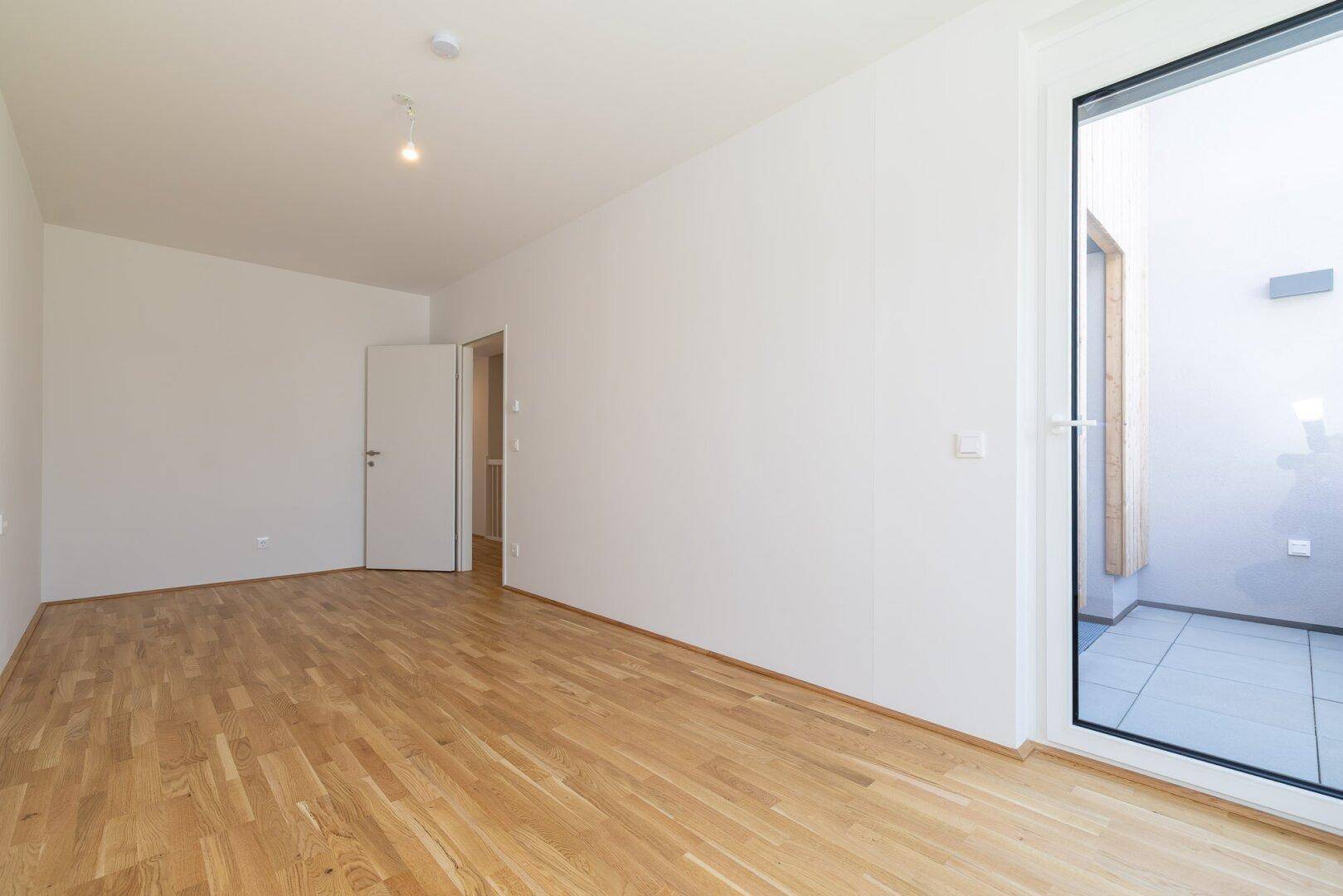 Helles Zimmer mit Parkettboden und Zugang zu einem gefliesten Außenbereich.