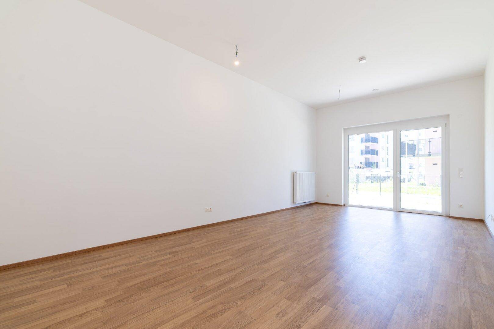 Heller, leerer Wohnraum mit Laminatboden, weißen Wänden und Zugang zur Terrasse.