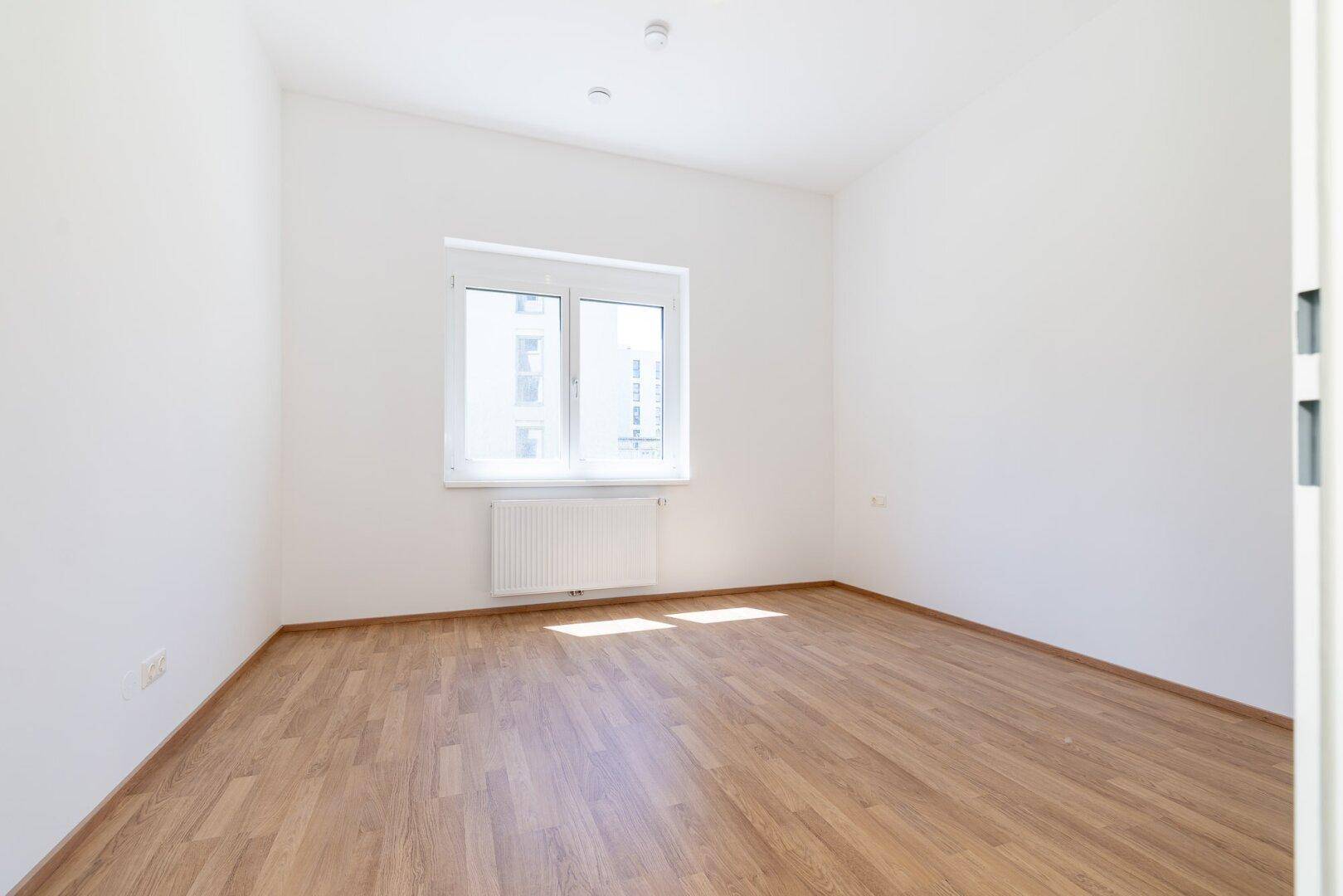 Helles Schlafzimmer mit Laminatboden, weiß gestrichenen Wänden und großem Fenster.
