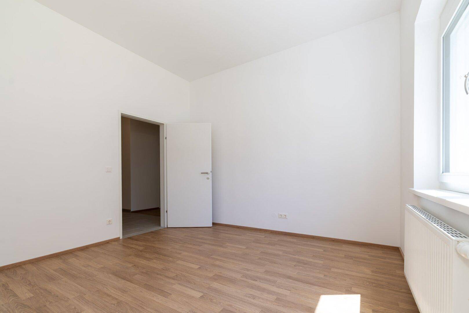 Leeres Schlafzimmer mit Laminatboden und offener Tür zu einem angrenzenden Raum.