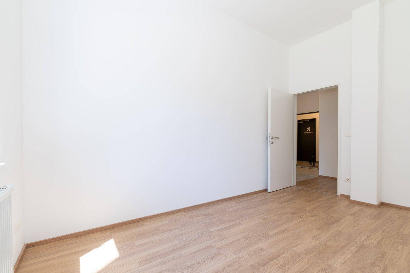 Leeres Schlafzimmer mit Laminatboden und offener Tür zu einem Flur oder Vorraum.