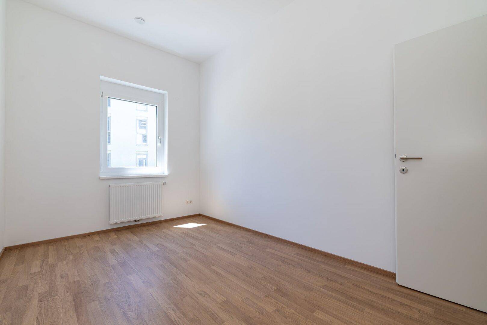 Unmöbliertes Schlafzimmer mit Laminatboden, weißer Wandfarbe und geschlossener Tür.
