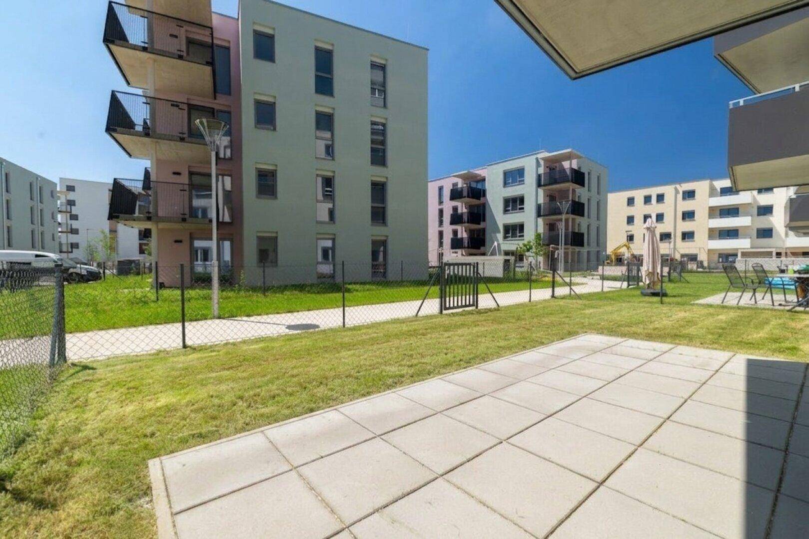Weitläufige Rasenfläche mit gepflastertem Bereich und farbenfrohen Neubauten im Hintergrund.