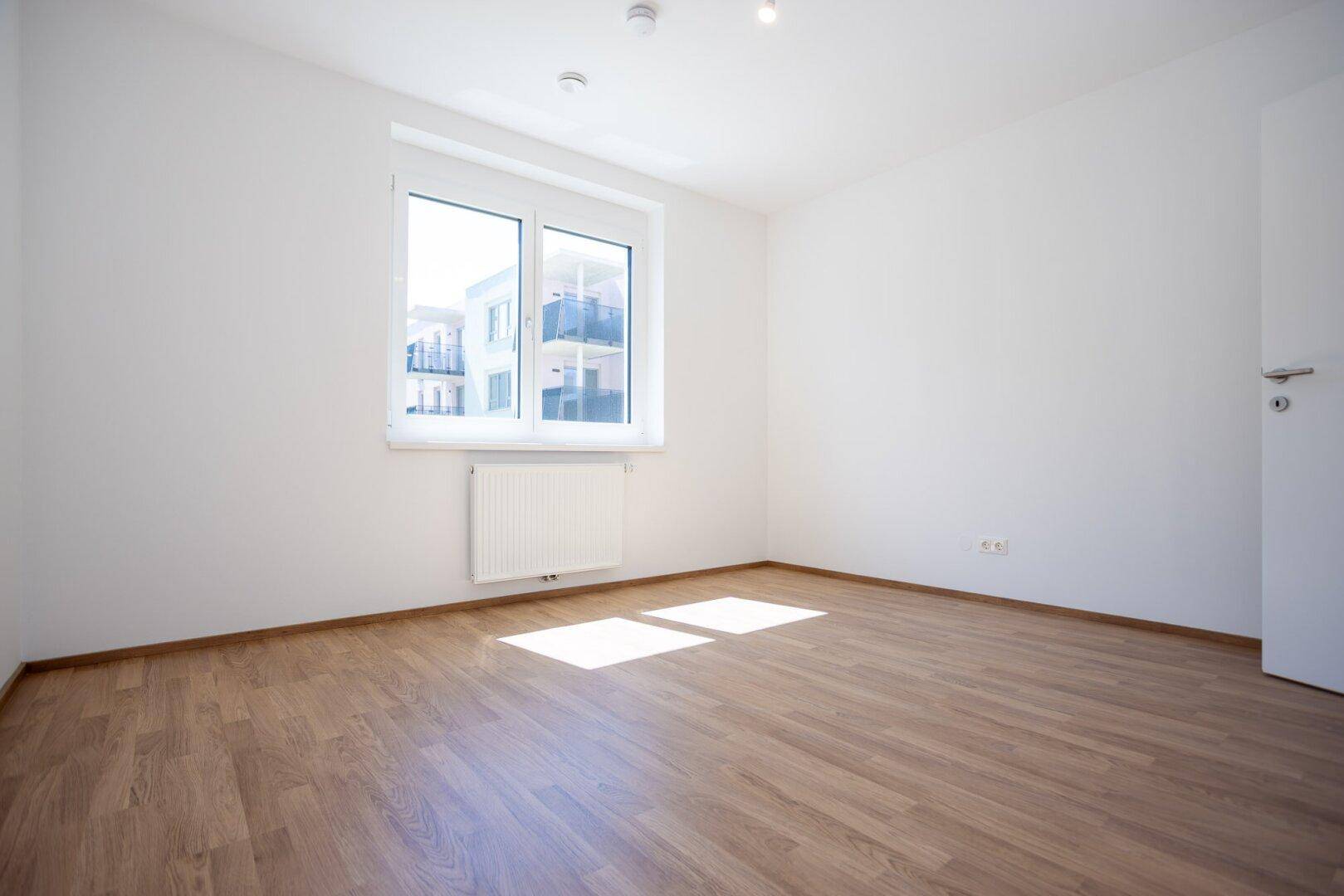 Lichtdurchflutetes Zimmer mit Laminatboden und Blick auf die moderne Wohnanlage.