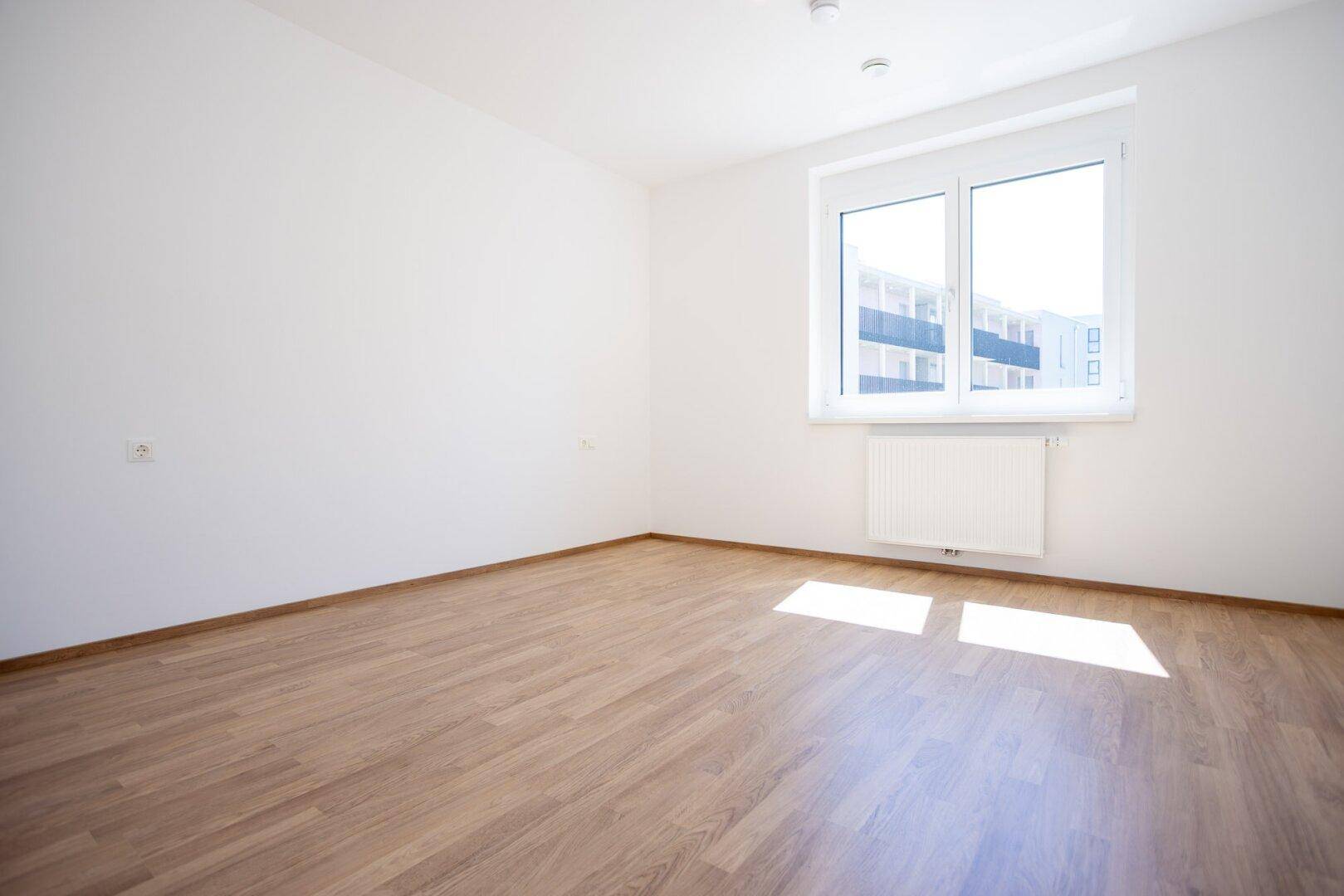 Helles, leeres Zimmer mit Laminatboden und großem Fenster, gut belichtet.