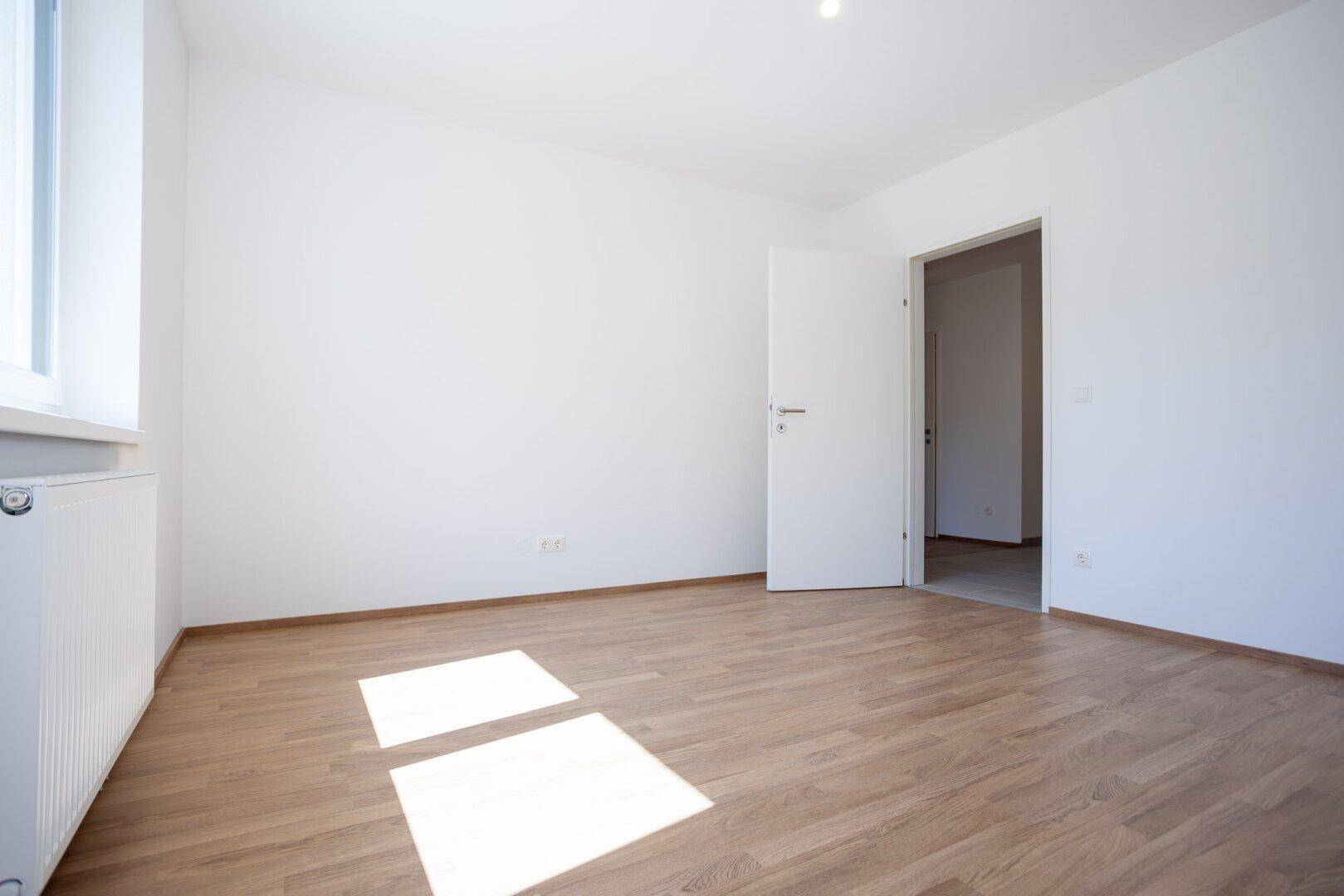 Geräumiges, helles Zimmer mit Laminatboden und offener Tür zu einem weiteren Raum.