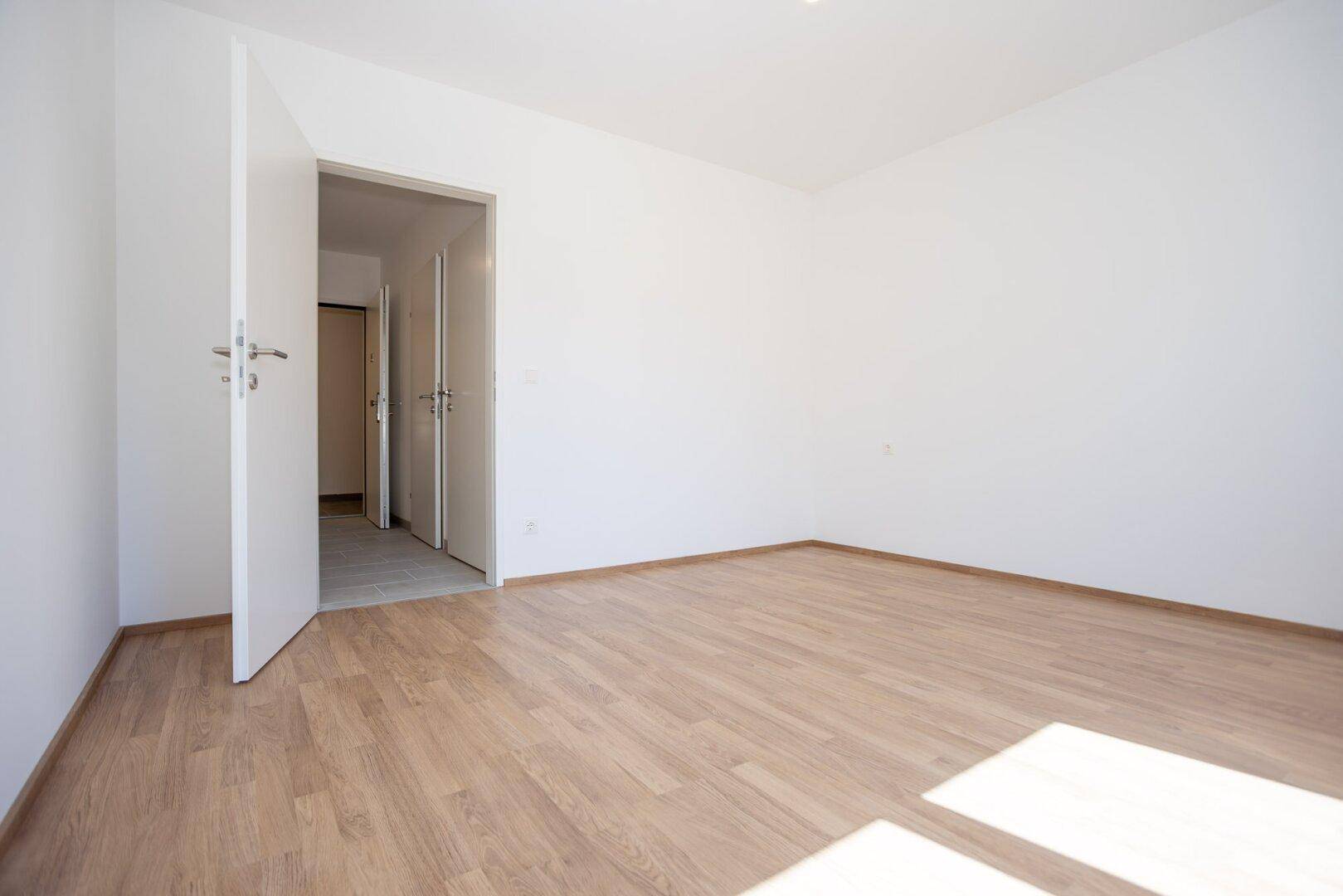 Leeres Zimmer mit Laminatboden und offener Tür, die in einen Flur führt.