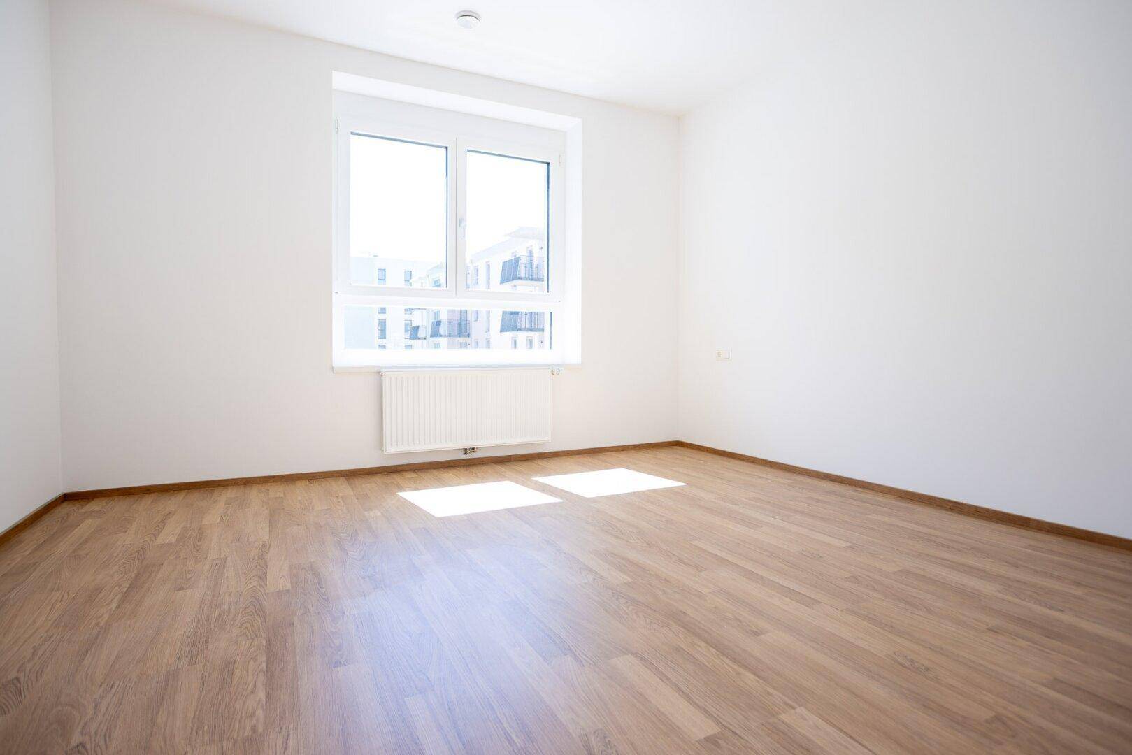 Helles Schlafzimmer mit Laminatboden und einem großen Fenster, das viel Sonnenlicht hereinlässt.